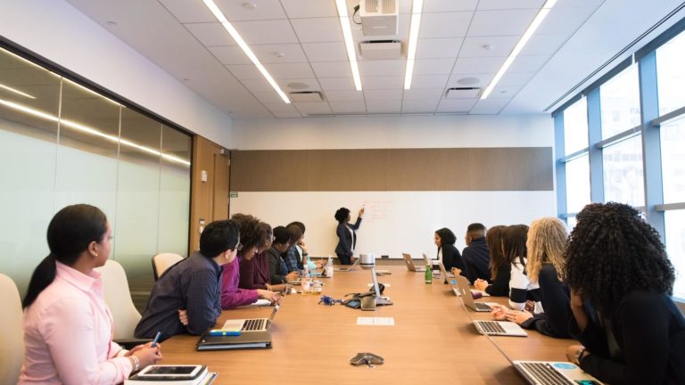 christina-wocintechchat-com-Q80LYxv_Tbs-unsplash(1)(1) Pessoas em mesa de reuniões estão de costas para a câmera olham para mulher apresentando dados em uma lousa branca no fundo da imagem. Ela é negra, tem cabelos crescos e aponta para a lousa