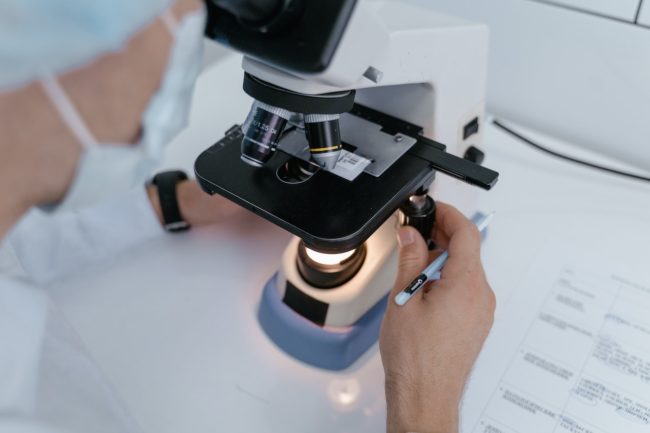 cientista de máscara e jaleco branco observa amostra em microscópio