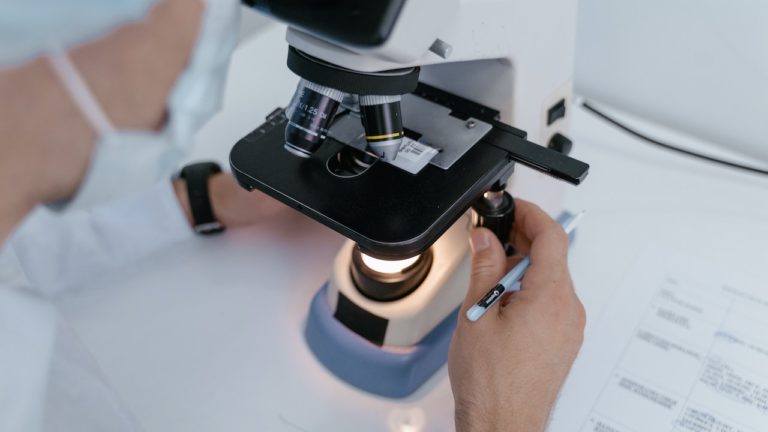 cientista de máscara e jaleco branco observa amostra em microscópio