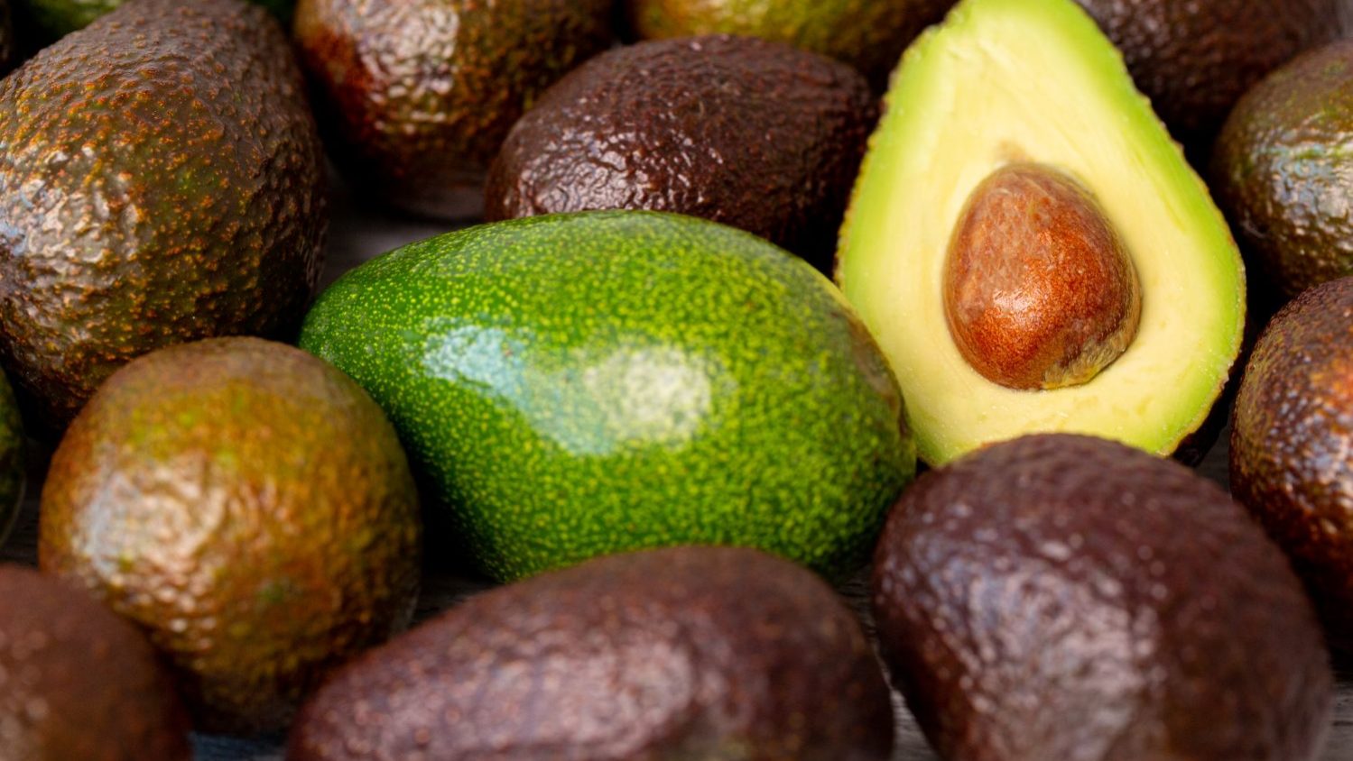 abacate-cortado-ao-lado-outro1 Abacates maduros agrupados, com um abacate cortado ao meio exibindo a polpa verde e o caroço.