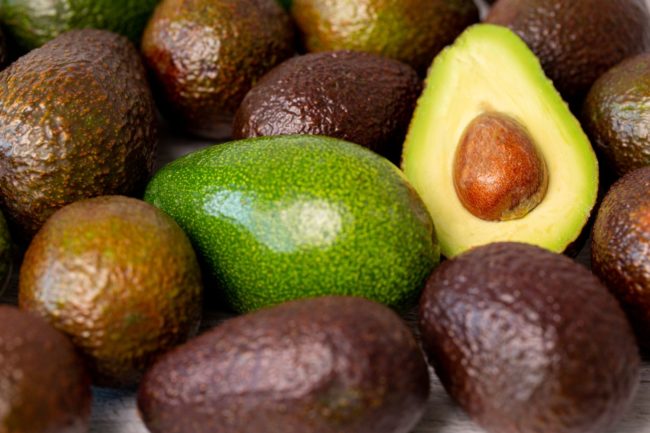abacate-cortado-ao-lado-outro1 Abacates maduros agrupados, com um abacate cortado ao meio exibindo a polpa verde e o caroço.