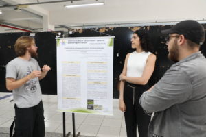 Bolsistas da Unifal-MG ao lado de pôster sobre divulgação científica apresentado no simpósio universitário.
