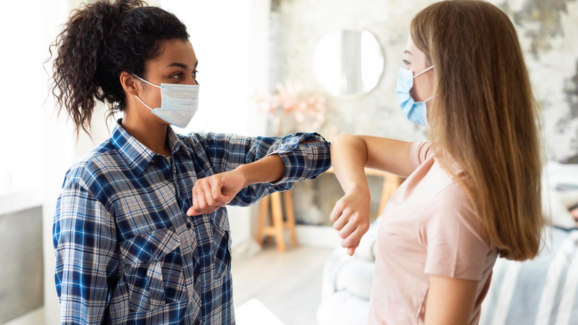 Duas mulheres de máscara se cumprimentam com os cotovelos durante a pandemia, simbolizando apoio e solidariedade.