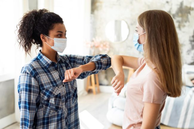 vista-lateral-de-amigas-com-mascaras-medicas-praticando-saudacao-de-cotovelo Duas mulheres de máscara se cumprimentam com os cotovelos durante a pandemia, simbolizando apoio e solidariedade.