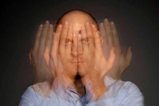Homem com expressão séria e mãos sobrepostas diante do rosto, sugerindo múltiplos estados mentais ou confusão, em fundo neutro.