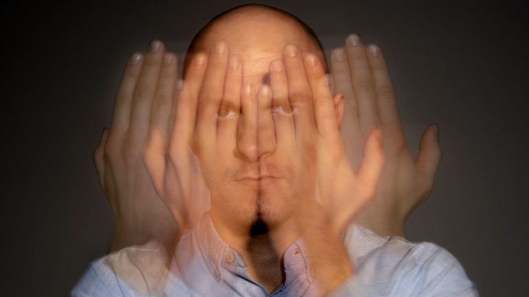 retrato-de-homem-que-sofre-de-esquizofrenia Homem com expressão séria e mãos sobrepostas diante do rosto, sugerindo múltiplos estados mentais ou confusão, em fundo neutro.