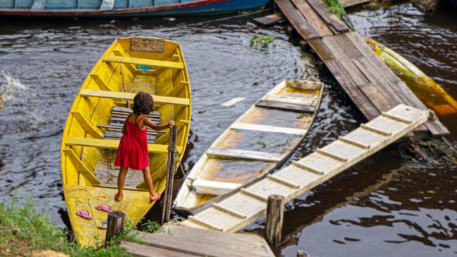 pexels-lu62-13557921 Criança em barco de madeira navegando em rio amazônico ao lado de passarelas e barcos.