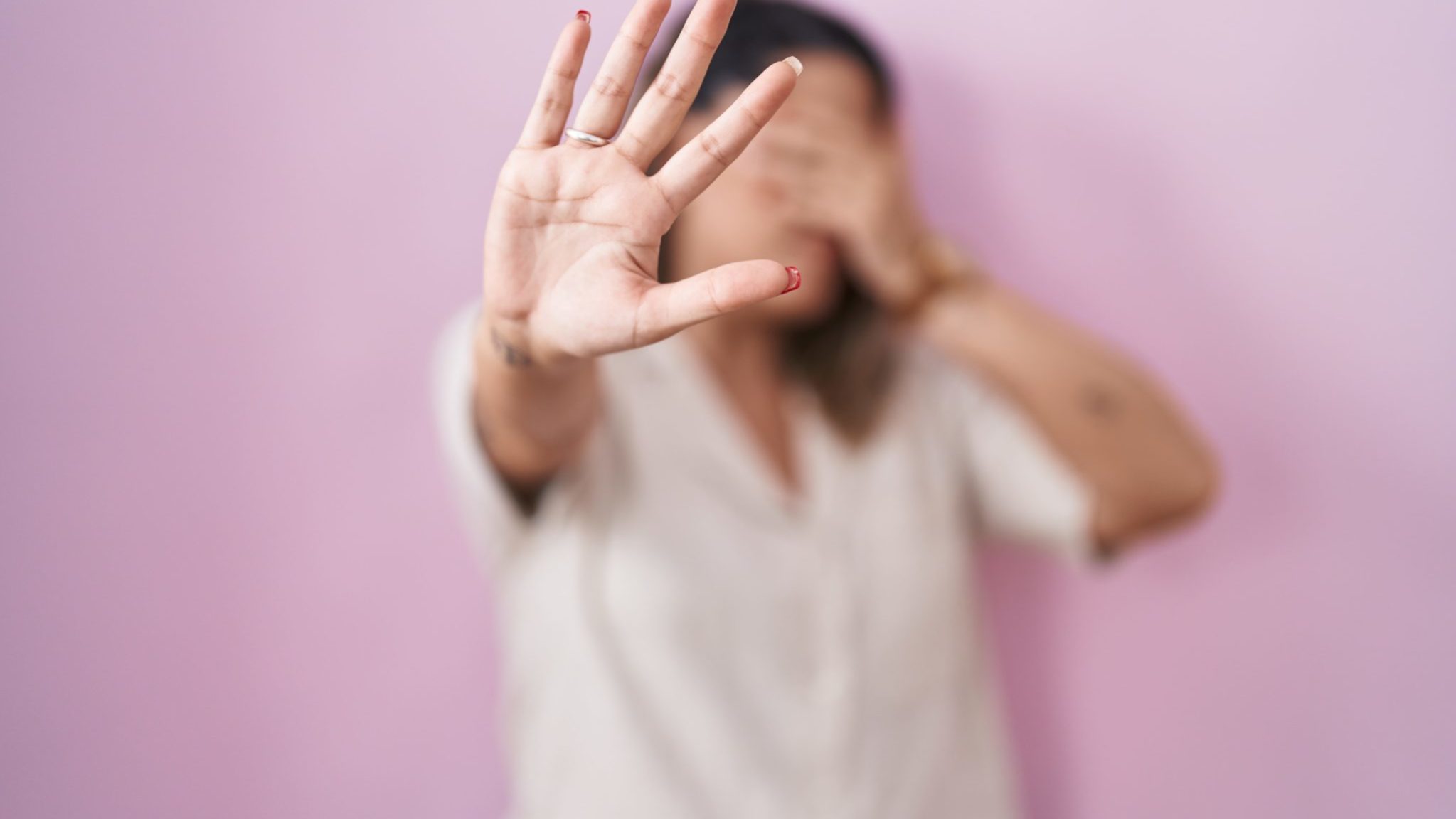Mulher de camisa clara, diante de parede rosa, cobre o rosto com uma mão e estende a outra em sinal de “pare”, representando resistência à violência doméstica.