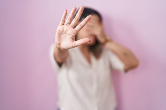 Mulher de camisa clara, diante de parede rosa, cobre o rosto com uma mão e estende a outra em sinal de “pare”, representando resistência à violência doméstica.