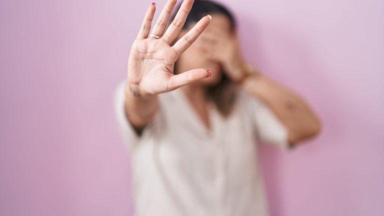 Mulher de camisa clara, diante de parede rosa, cobre o rosto com uma mão e estende a outra em sinal de “pare”, representando resistência à violência doméstica.