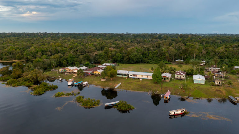 Casas ribeirinhas de madeira à beira d’água na Reserva Amanã, no Amazonas, cercadas por vegetação e pequenas embarcações, retratando o cotidiano das comunidades que lidam com o acúmulo de resíduos plásticos.
