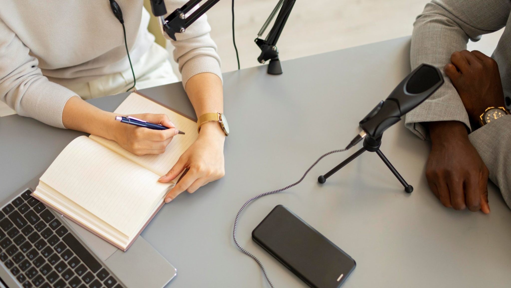 mulher-fazendo-perguntas-em-um-podcast Duas pessoas sentadas à mesa diante de microfones; uma anota em um caderno enquanto a outra mantém os braços apoiados, sugerindo uma situação de entrevista