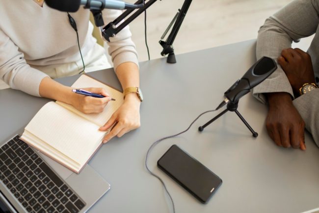 Duas pessoas sentadas à mesa diante de microfones; uma anota em um caderno enquanto a outra mantém os braços apoiados, sugerindo uma situação de entrevista
