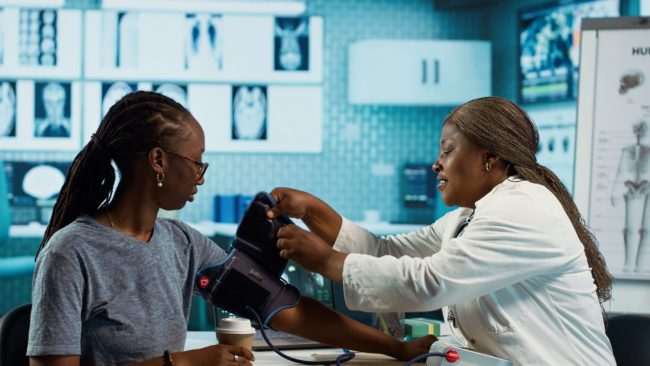 black-medic-practitioner-takes-blood-pressure-with-blood-pressure-monitor Profissional de saúde mede a pressão arterial de uma mulher em atendimento clínico, usando aparelho de aferição no braço.